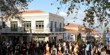 Σέρβια 28ης Οκτωβρίου: Χορευτές σε παραδοσιακές στολές χορεύουν στην Κεντρική Πλατεία των Σερβίων με θεατές και ελληνικές σημαίες στο φόντο.