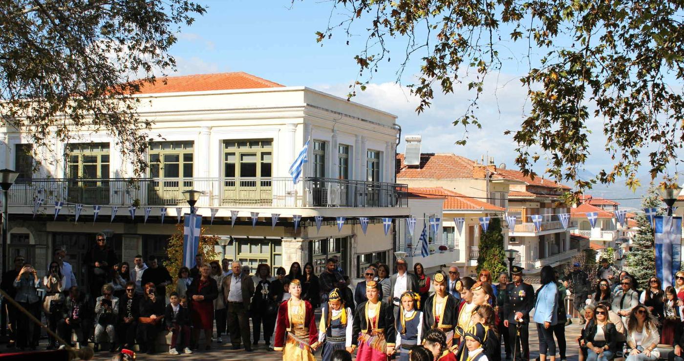 Σέρβια 28ης Οκτωβρίου: Χορευτές σε παραδοσιακές στολές χορεύουν στην Κεντρική Πλατεία των Σερβίων με θεατές και ελληνικές σημαίες στο φόντο.