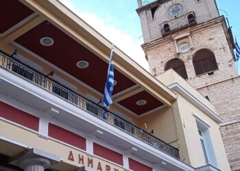 Το Δημαρχείο της Κοζάνης με τον πύργο του ρολογιού και την ελληνική σημαία, συμβολίζοντας την κατάργηση των πιστοποιητικών κοζάνης και την αυτεπάγγελτη αναζήτηση.