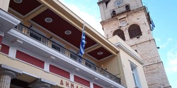 Το Δημαρχείο της Κοζάνης με τον πύργο του ρολογιού και την ελληνική σημαία, συμβολίζοντας την κατάργηση των πιστοποιητικών κοζάνης και την αυτεπάγγελτη αναζήτηση.