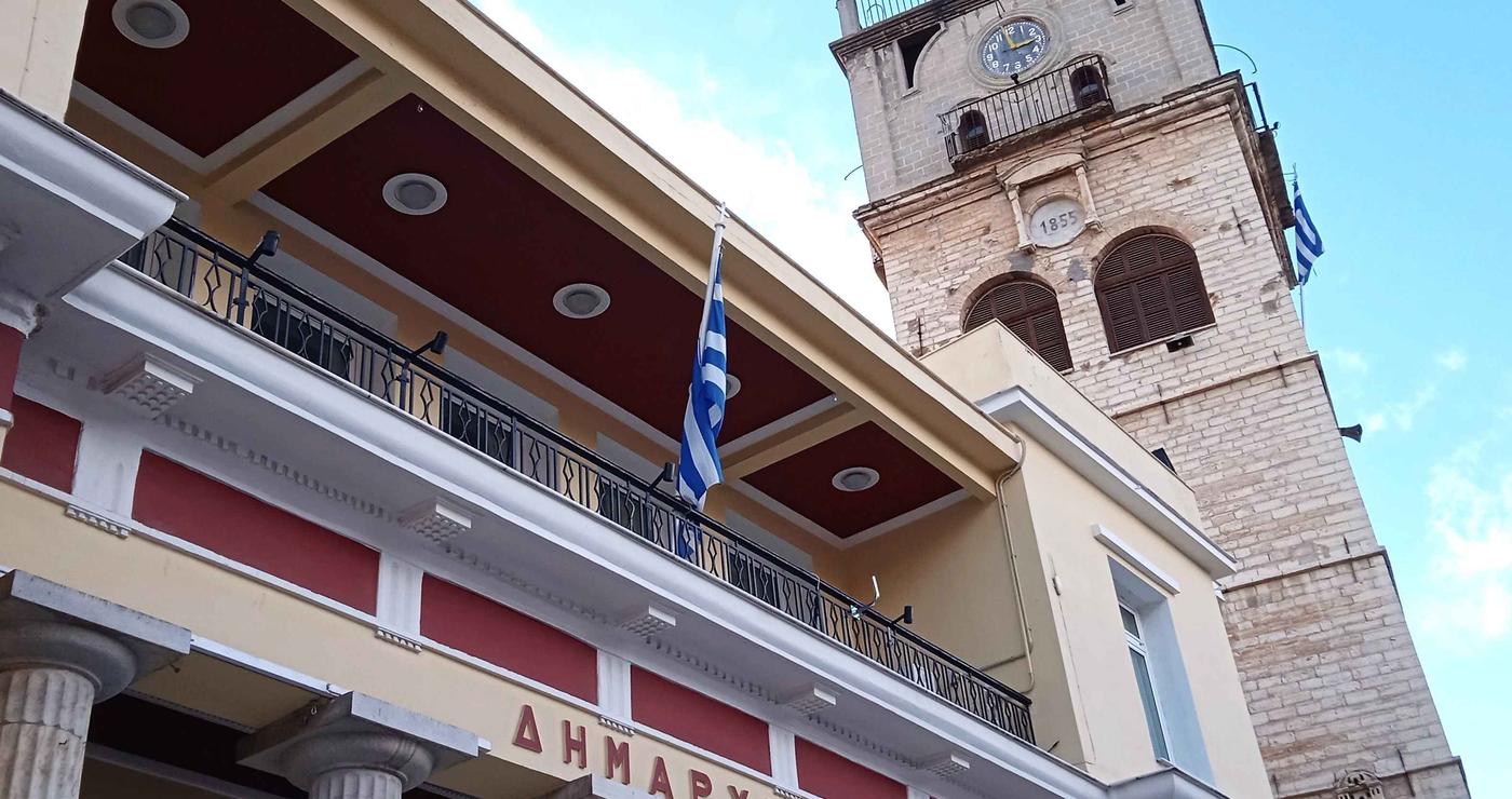 Το Δημαρχείο της Κοζάνης με τον πύργο του ρολογιού και την ελληνική σημαία, συμβολίζοντας την κατάργηση των πιστοποιητικών κοζάνης και την αυτεπάγγελτη αναζήτηση.