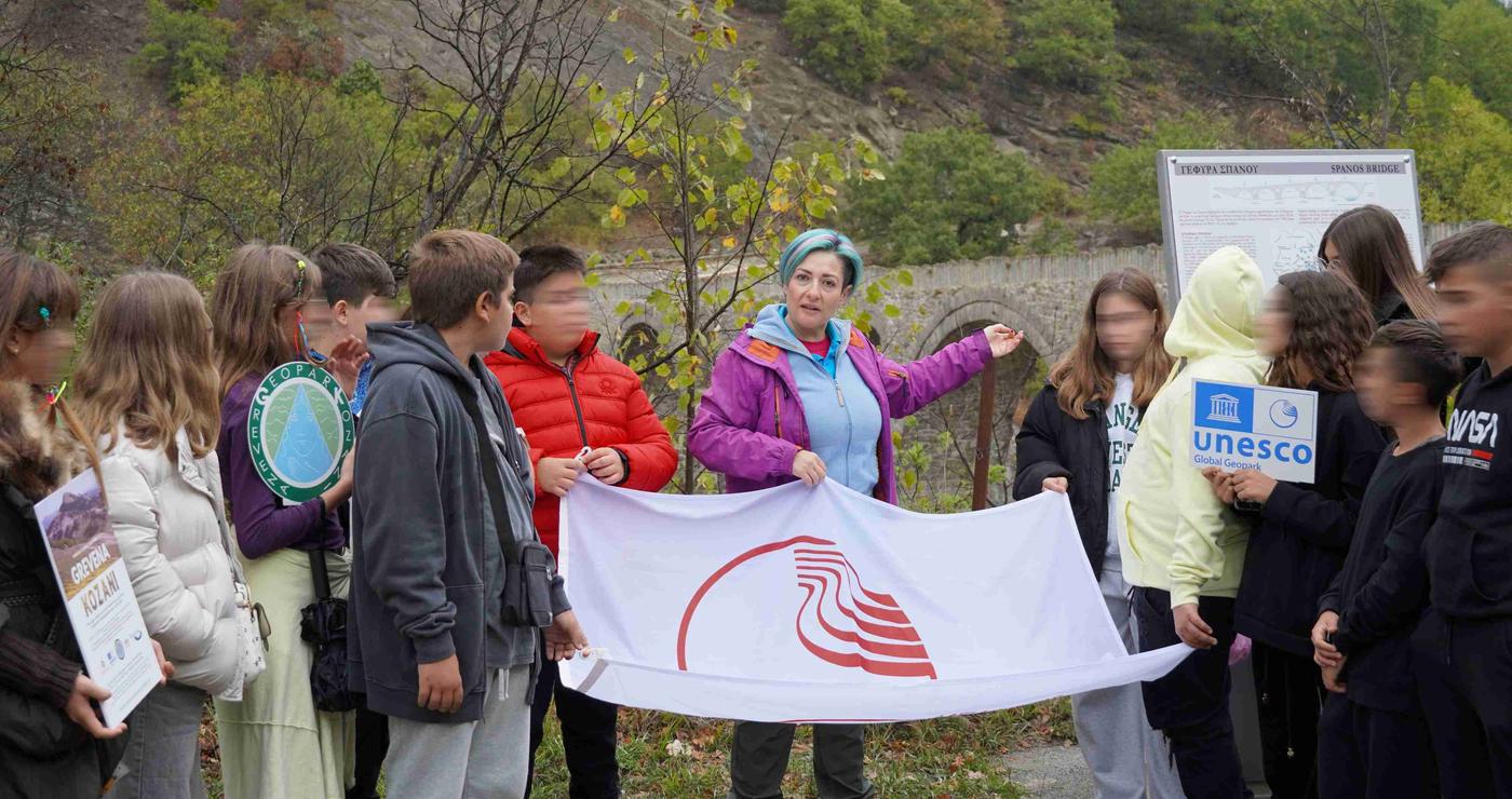 Μια ομάδα μαθητών του Μουσικού Γυμνασίου Βαρθολομιού ακούει με προσοχή μια γυναίκα που τους παρουσιάζει το Γεωπάρκο Γρεβενών-Κοζάνης, κρατώντας μια σημαία με το λογότυπο του UNESCO.