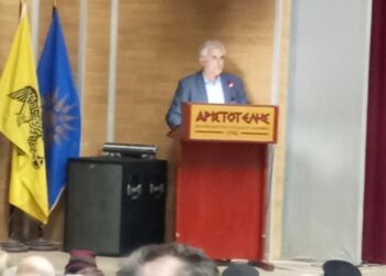 Ένας άνδρας μιλάει σε ένα κοινό στην παράσταση του θεατρικού εργαστηρίου Φλώρινας, με την ελληνική και την σημαία της Μητροπόλεως Φλωρίνης να δεσπόζουν στο βάθος.