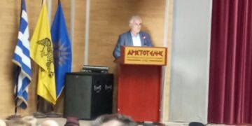 Ένας άνδρας μιλάει σε ένα κοινό στην παράσταση του θεατρικού εργαστηρίου Φλώρινας, με την ελληνική και την σημαία της Μητροπόλεως Φλωρίνης να δεσπόζουν στο βάθος.