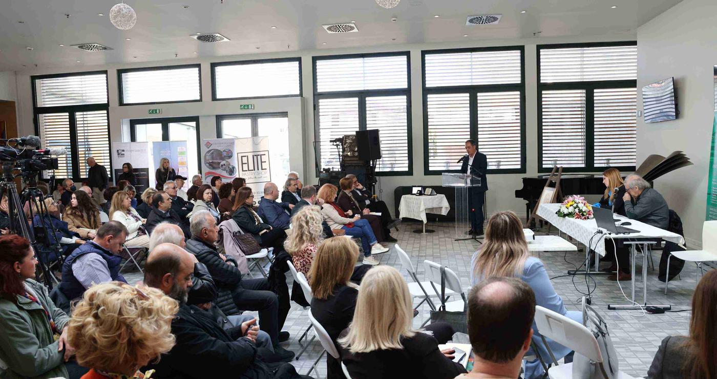 Ένα πλήθος ανθρώπων παρακολουθεί ένα συμπόσιο λογοτεχνίας σε ένα ευρύχωρο δωμάτιο με μεγάλα παράθυρα στην Κοζάνη.