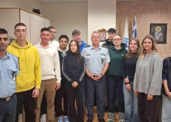 Μια ομάδα δόκιμων αστυφυλάκων στέκεται χαμογελαστοί σε έναν εσωτερικό χώρο, φορώντας καθημερινά ρούχα και στολές, με ελληνική σημαία στο βάθος, κατά την κατάταξή τους στη Δυτική Μακεδονία.