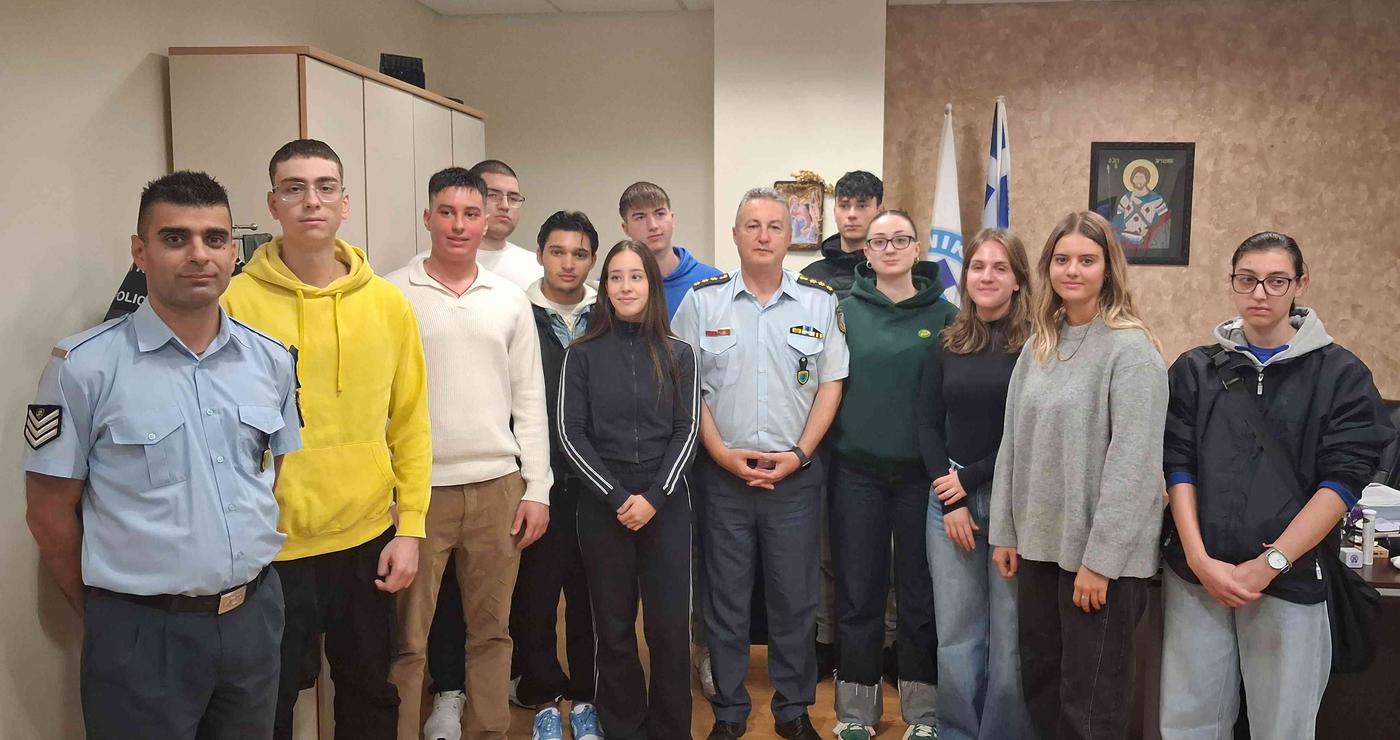 Μια ομάδα δόκιμων αστυφυλάκων στέκεται χαμογελαστοί σε έναν εσωτερικό χώρο, φορώντας καθημερινά ρούχα και στολές, με ελληνική σημαία στο βάθος, κατά την κατάταξή τους στη Δυτική Μακεδονία.