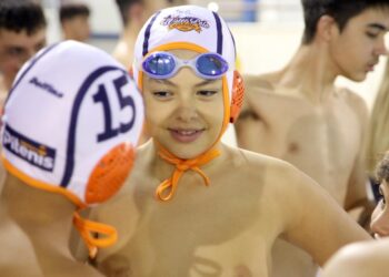 Ένας νεαρός αθλητής Water Polo Κοζάνης χαμογελά στον φακό φορώντας σκουφάκι και γυαλιά κολύμβησης, περιτριγυρισμένος από τους συμπαίκτες του στο κολυμβητήριο.