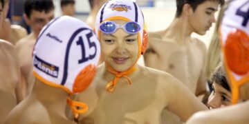 Ένας νεαρός αθλητής Water Polo Κοζάνης χαμογελά στον φακό φορώντας σκουφάκι και γυαλιά κολύμβησης, περιτριγυρισμένος από τους συμπαίκτες του στο κολυμβητήριο.