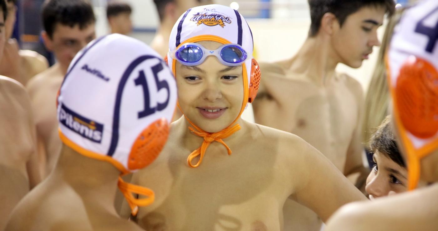 Ένας νεαρός αθλητής Water Polo Κοζάνης χαμογελά στον φακό φορώντας σκουφάκι και γυαλιά κολύμβησης, περιτριγυρισμένος από τους συμπαίκτες του στο κολυμβητήριο.