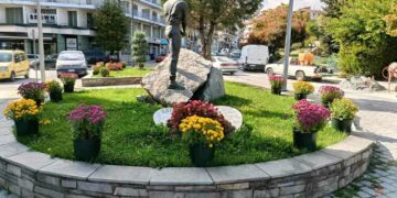 Ένας ανδρικός μνημειακός αγάλμα βρίσκεται στο κέντρο ενός κυκλικού κηπάριου γεμάτου εποχιακά λουλούδια, μέρος των ανθοφυτεύσεων Φλώρινας, με κτίρια και αυτοκίνητα στο φόντο.