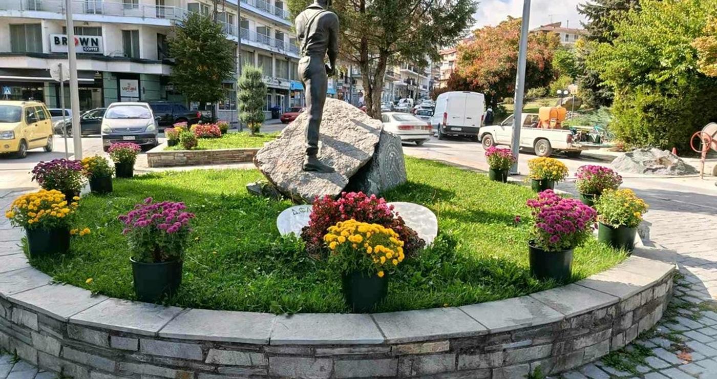 Ένας ανδρικός μνημειακός αγάλμα βρίσκεται στο κέντρο ενός κυκλικού κηπάριου γεμάτου εποχιακά λουλούδια, μέρος των ανθοφυτεύσεων Φλώρινας, με κτίρια και αυτοκίνητα στο φόντο.