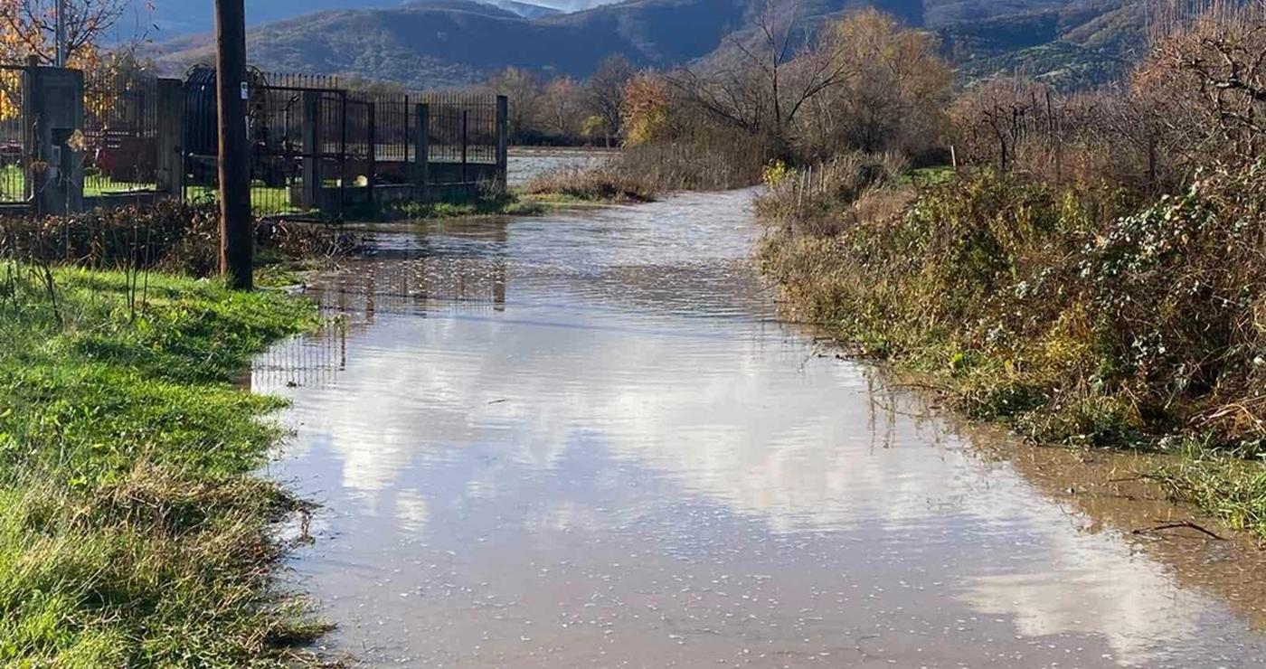 A flooded rural road in Florina, with mountains visible in the background, showcasing the extent of the Πλημμύρες Φλώρινας.
