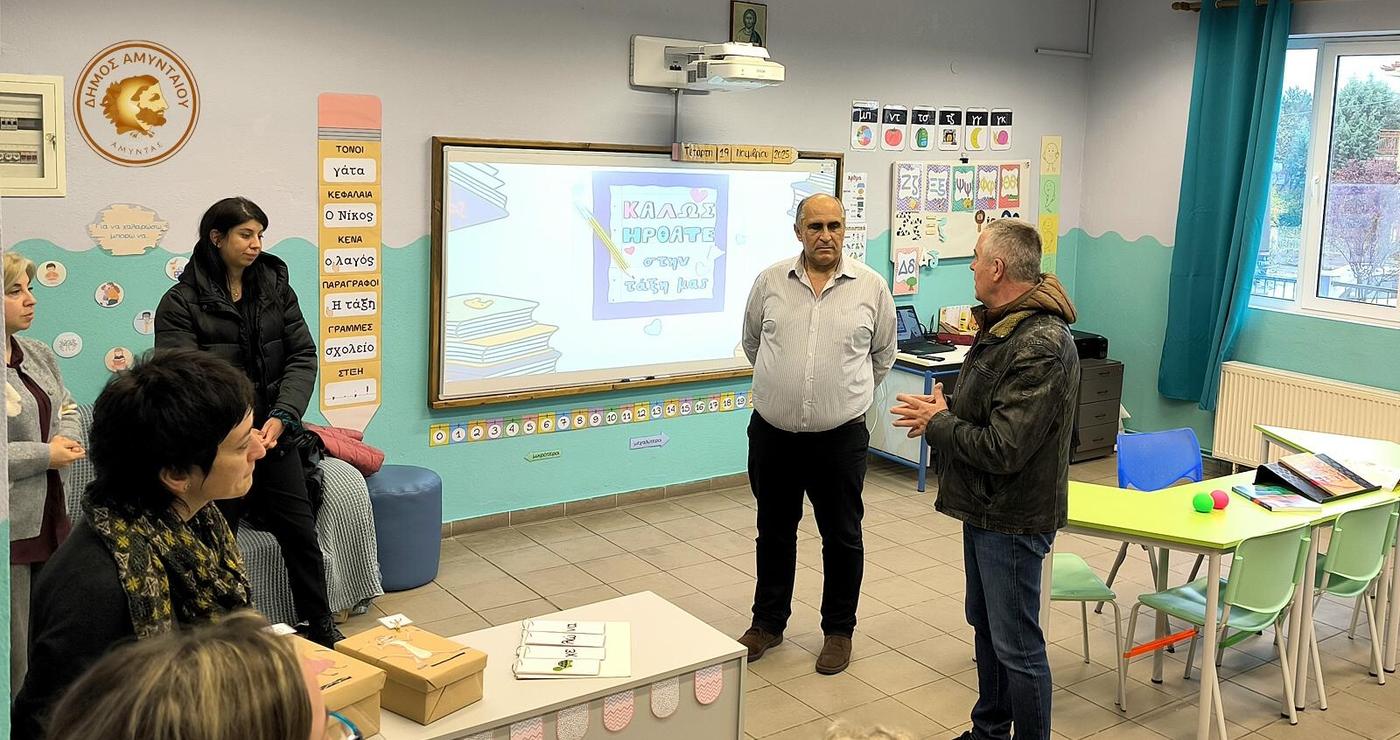 Μια δασκάλα, ένας άνδρας και μια ομάδα μαθητών συζητούν στο νέο Τμήμα Ένταξης Αμυνταίου, με τον άνδρα να παρουσιάζει κάτι με ενθουσιασμό.