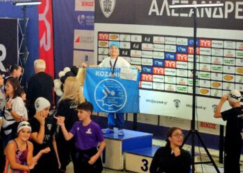 Μια ομάδα αθλητών της Κολυμβητικής Κοζάνης στέκεται περήφανα με την σημαία της ομάδας τους δίπλα στην πισίνα, μετά την επιτυχημένη συμμετοχή τους στους αγώνες «Αλεξάνδρεια Junior» στη Θεσσαλονίκη.