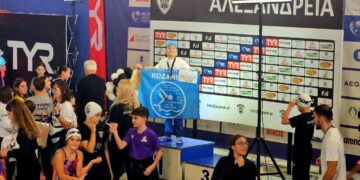 Μια ομάδα αθλητών της Κολυμβητικής Κοζάνης στέκεται περήφανα με την σημαία της ομάδας τους δίπλα στην πισίνα, μετά την επιτυχημένη συμμετοχή τους στους αγώνες «Αλεξάνδρεια Junior» στη Θεσσαλονίκη.