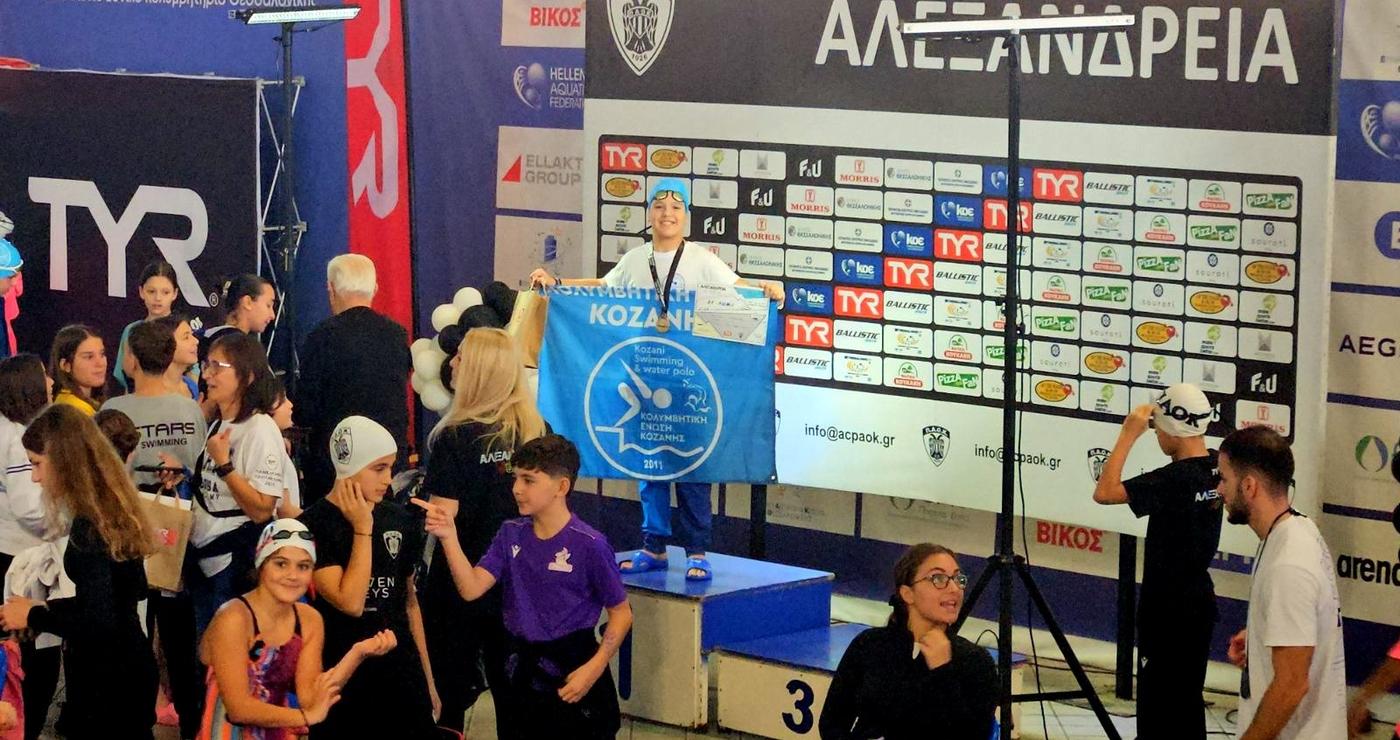 Μια ομάδα αθλητών της Κολυμβητικής Κοζάνης στέκεται περήφανα με την σημαία της ομάδας τους δίπλα στην πισίνα, μετά την επιτυχημένη συμμετοχή τους στους αγώνες «Αλεξάνδρεια Junior» στη Θεσσαλονίκη.