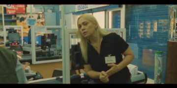 Μια γυναίκα με ξανθά μαλλιά στέκεται σε ένα ταμείο ενός σούπερ μάρκετ, κοιτάζοντας προς τα δεξιά, μέρος της ταινίας Super Story Market.