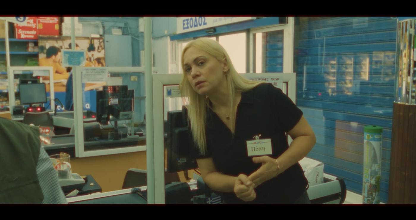 Μια γυναίκα με ξανθά μαλλιά στέκεται σε ένα ταμείο ενός σούπερ μάρκετ, κοιτάζοντας προς τα δεξιά, μέρος της ταινίας Super Story Market.