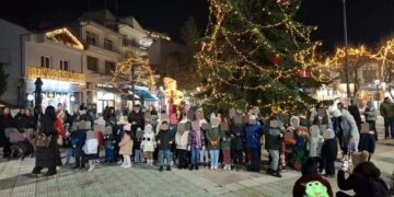 Πλήθος παιδιών και γονέων συγκεντρώνονται γύρω από ένα φωτισμένο χριστουγεννιάτικο δέντρο στην Γιορτή Ζαχαρωτών στη Φλώρινα.