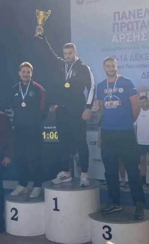 Ο Θωμάς Γκιουλέκας στέκεται στο πρώτο σκαλί του βάθρου φορώντας ένα χρυσό μετάλλιο και με χαμόγελο, έχοντας κατακτήσει την πρώτη θέση στην άρση βαρών.