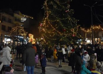 Ένα πλήθος ανθρώπων συγκεντρώνεται γύρω από το φωτισμένο χριστουγεννιάτικο δέντρο στην πλατεία της Φλώρινας κατά τη διάρκεια των χριστουγεννιάτικων εκδηλώσεων.