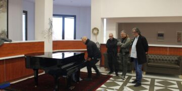 Άνδρες παρακολουθούν έναν άνδρα να στήνει το νέο πιάνο στο Δημοτικό Ωδείο Σερβίων, μια σημαντική προσθήκη για τη μουσική εκπαίδευση.