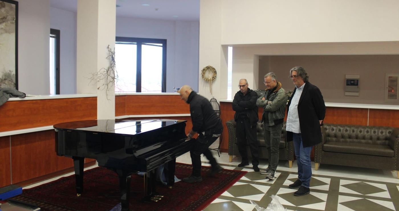 Άνδρες παρακολουθούν έναν άνδρα να στήνει το νέο πιάνο στο Δημοτικό Ωδείο Σερβίων, μια σημαντική προσθήκη για τη μουσική εκπαίδευση.