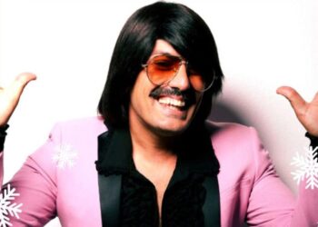 Ο Τόνι Σφήνος ποζάρει χαμογελαστός για την Καστοριά Πρωτοχρονιά, φορώντας ένα ροζ σακάκι και με χιονονιφάδες γύρω του.