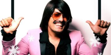 Ο Τόνι Σφήνος ποζάρει χαμογελαστός για την Καστοριά Πρωτοχρονιά, φορώντας ένα ροζ σακάκι και με χιονονιφάδες γύρω του.