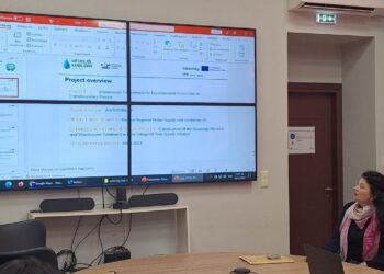 Ένας άνδρας μιλάει σε μια συνάντηση για το έργο WaterTrap, με την παρουσίαση να προβάλλεται στην οθόνη και άλλους συμμετέχοντες στο τραπέζι, εστιάζοντας στην επεξεργασία λυμάτων στην Πρέσπα.