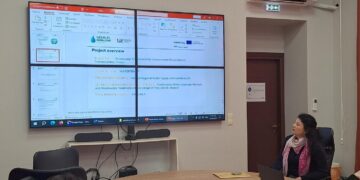 Ένας άνδρας μιλάει σε μια συνάντηση για το έργο WaterTrap, με την παρουσίαση να προβάλλεται στην οθόνη και άλλους συμμετέχοντες στο τραπέζι, εστιάζοντας στην επεξεργασία λυμάτων στην Πρέσπα.