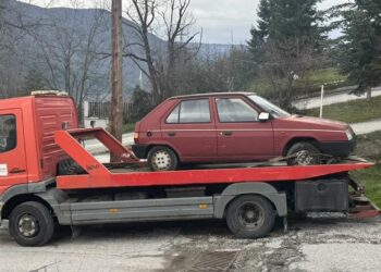 Ένα κόκκινο φορτηγό μεταφέρει ένα εγκαταλελειμμένο λευκό αυτοκίνητο δίπλα σε έναν δρόμο στη Φλώρινα.