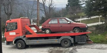 Ένα κόκκινο φορτηγό μεταφέρει ένα εγκαταλελειμμένο λευκό αυτοκίνητο δίπλα σε έναν δρόμο στη Φλώρινα.