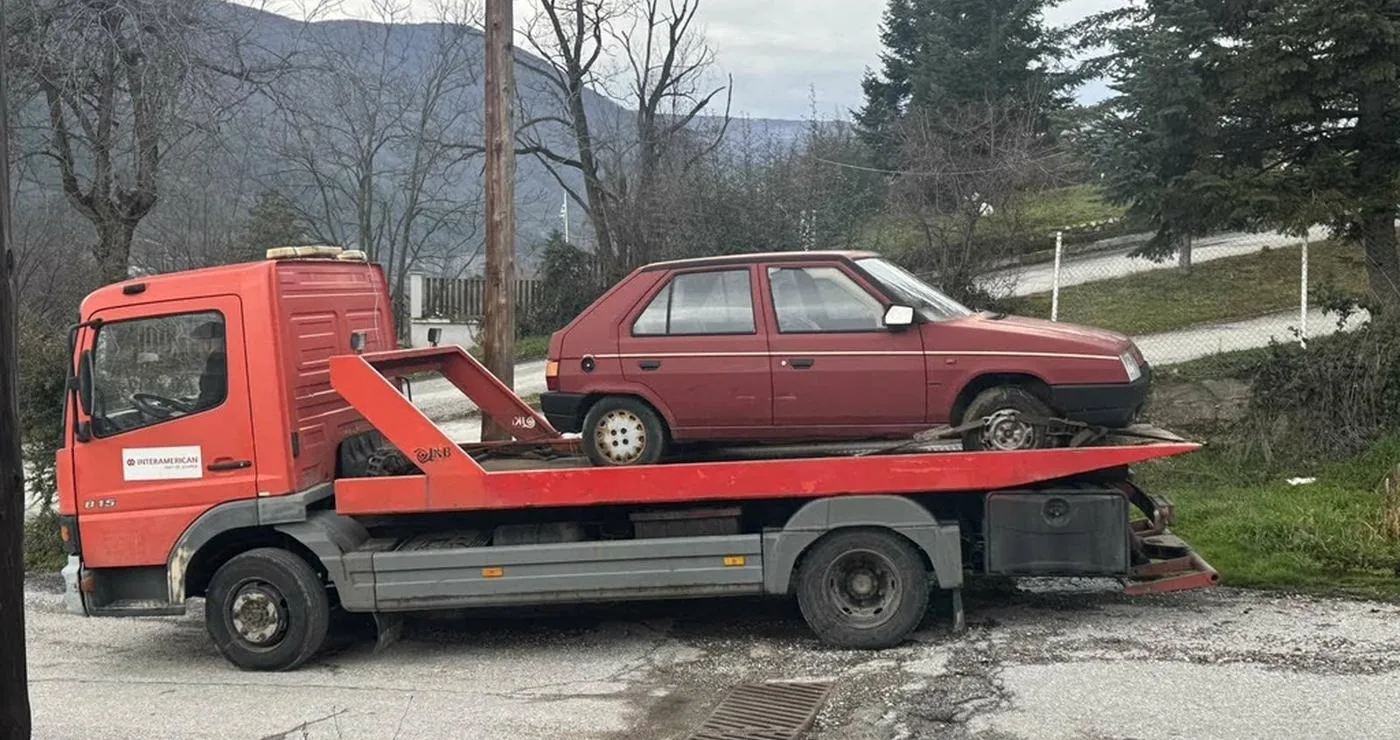 Ένα κόκκινο φορτηγό μεταφέρει ένα εγκαταλελειμμένο λευκό αυτοκίνητο δίπλα σε έναν δρόμο στη Φλώρινα.