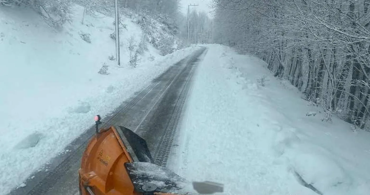 Ένα εκχιονιστικό μηχάνημα αποχιονίζει έναν δρόμο καλυμμένο με χιόνι στη Φλώρινα, με δέντρα και στις δύο πλευρές.