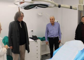 Μια γυναίκα γιατρός εξηγεί σε τρεις άνδρες τις νέες υποδομές του ανακαινισμένου Κέντρου Υγείας Σερβίων, ενώ βρίσκονται σε ένα καθαρό εξεταστήριο με ένα μεγάλο φωτιστικό πάνω από το κρεβάτι.