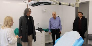 Μια γυναίκα γιατρός εξηγεί σε τρεις άνδρες τις νέες υποδομές του ανακαινισμένου Κέντρου Υγείας Σερβίων, ενώ βρίσκονται σε ένα καθαρό εξεταστήριο με ένα μεγάλο φωτιστικό πάνω από το κρεβάτι.