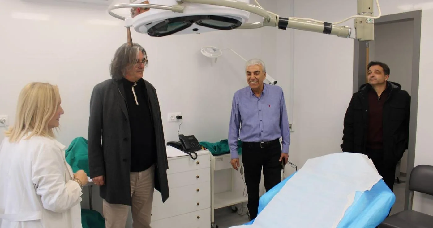 Μια γυναίκα γιατρός εξηγεί σε τρεις άνδρες τις νέες υποδομές του ανακαινισμένου Κέντρου Υγείας Σερβίων, ενώ βρίσκονται σε ένα καθαρό εξεταστήριο με ένα μεγάλο φωτιστικό πάνω από το κρεβάτι.