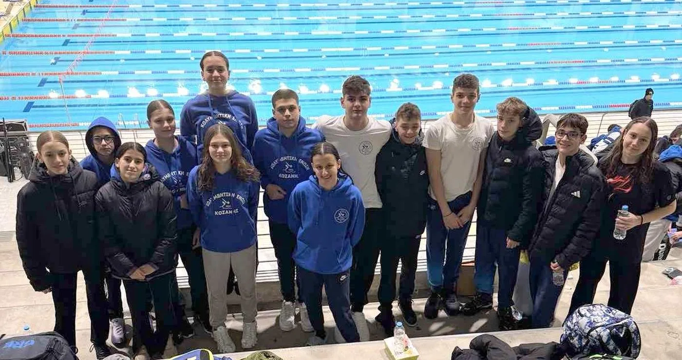 Η ομάδα κολύμβησης της Κοζάνης στέκεται περήφανα δίπλα στην πισίνα στην Καβάλα, φορώντας τα μπλε τους φλιτζάνια με το λογότυπο, μετά τους αγώνες κολύμβησης.