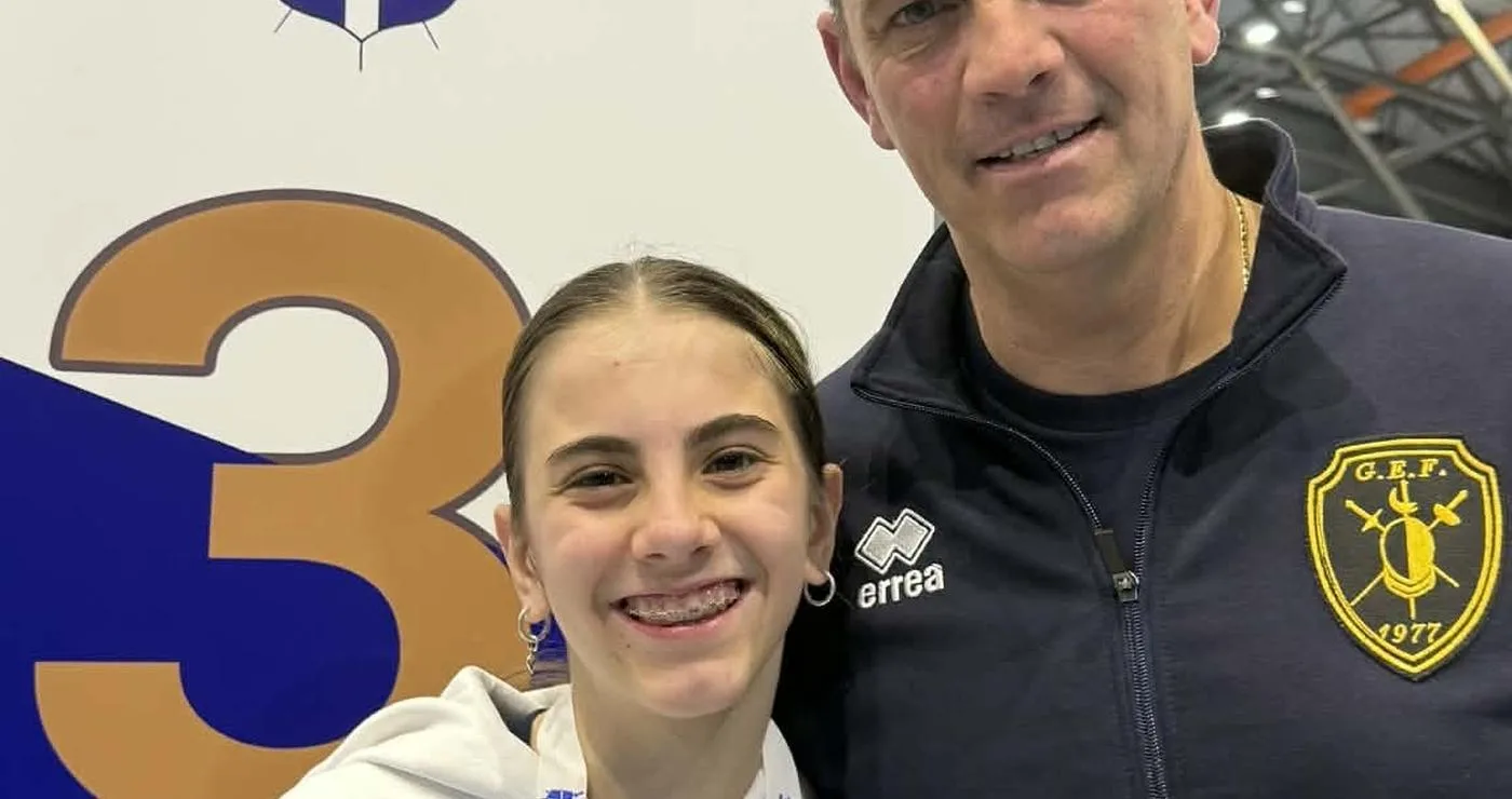 Η Πανδώρα Γουσοπούλου, μια νεαρή αθλήτρια της ξιφασκίας, χαμογελά δίπλα στον προπονητή της, φορώντας το χάλκινο μετάλλιο της στο Πανελλήνιο Κύπελλο, επιτυγχάνοντας τη Γουσοπούλου στο βάθρο.