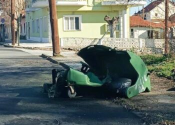 Ένας πράσινος κάδος απορριμμάτων Φλώρινας με ανοιχτό το καπάκι βρίσκεται σε δρόμο με σπίτια και δέντρα, δείχνοντας την ανάγκη προσοχής στη στάχτη.