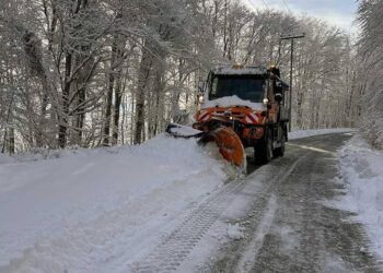 Ένα πορτοκαλί μηχάνημα αποχιονισμού καθαρίζει ένα δρόμο καλυμμένο με χιόνι, ανάμεσα σε δέντρα με παγωμένα κλαδιά στην Φλώρινα, κατά τη διάρκεια του αποχιονισμού.