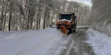 Ένα πορτοκαλί μηχάνημα αποχιονισμού καθαρίζει ένα δρόμο καλυμμένο με χιόνι, ανάμεσα σε δέντρα με παγωμένα κλαδιά στην Φλώρινα, κατά τη διάρκεια του αποχιονισμού.
