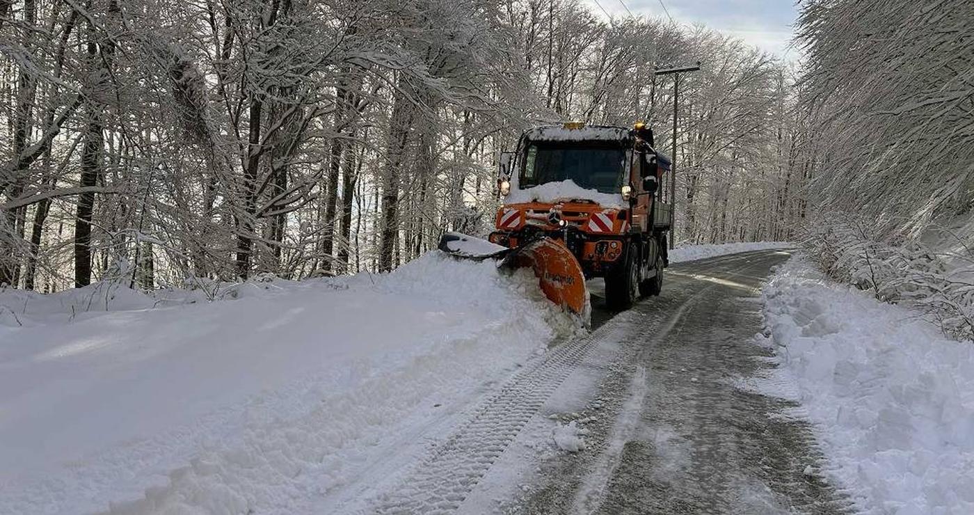 Ένα πορτοκαλί μηχάνημα αποχιονισμού καθαρίζει ένα δρόμο καλυμμένο με χιόνι, ανάμεσα σε δέντρα με παγωμένα κλαδιά στην Φλώρινα, κατά τη διάρκεια του αποχιονισμού.