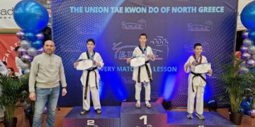 Three young taekwondo athletes stand on a podium at the Taekwondo Northern Greece championship, proudly holding their certificates and wearing black belts.