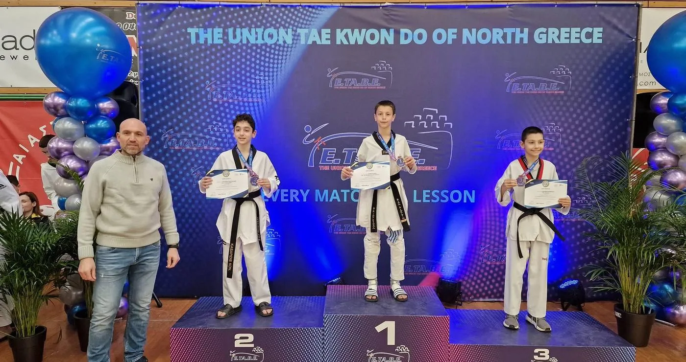 Three young taekwondo athletes stand on a podium at the Taekwondo Northern Greece championship, proudly holding their certificates and wearing black belts.