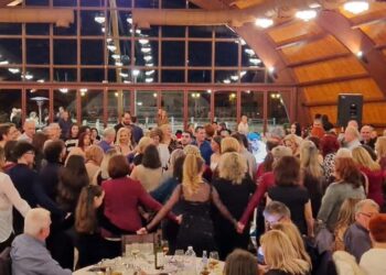 Ένα πλήθος ανθρώπων χορεύουν σε κύκλο στον ετήσιο χορό Κοζάνης στο Event Plaza, με τραπέζια και καλεσμένους να τους παρακολουθούν.