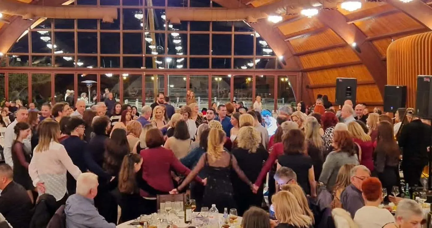 Ένα πλήθος ανθρώπων χορεύουν σε κύκλο στον ετήσιο χορό Κοζάνης στο Event Plaza, με τραπέζια και καλεσμένους να τους παρακολουθούν.