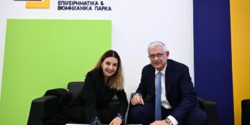Two people, a woman and a man, are signing a document at a table during the ΔΥΠΑ – ΕΤΒΑ ΒΙ.ΠΕ. cooperation agreement signing event.